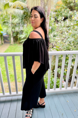 Side view of Bianca in the Voyager Jumpsuit in Black knit, cape worn off the shoulders, hand in the side pocket — Libertine The Label