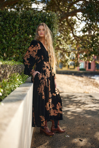 Woman in a black and gold floral dress standing outdoors with greenery in the background