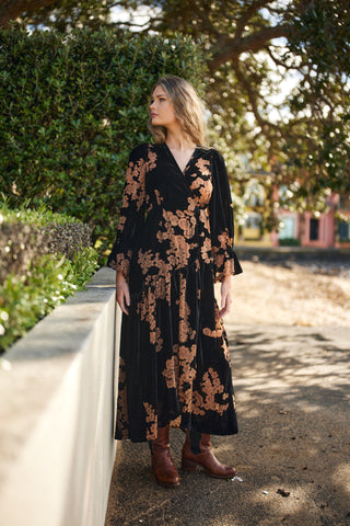 Woman in a black and brown floral dress standing outdoors with greenery in the background