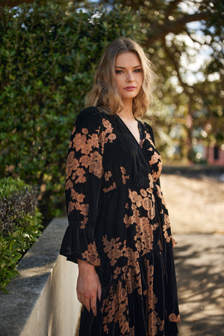 Woman wearing a black floral dress standing outdoors with greenery in the background