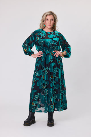 Woman wearing a teal orchid velvet dress, featuring a floral design and ankle boots, showcasing a stylish and elegant look.