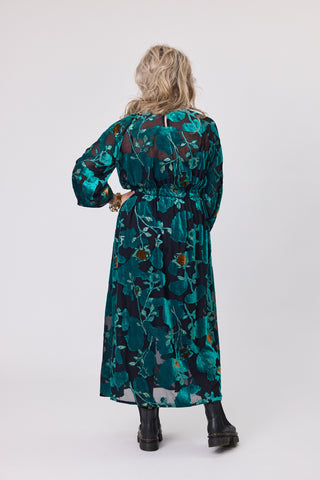 Back view of a woman wearing a teal orchid velvet dress with floral design, showcasing its flowy skirt and relaxed fit.