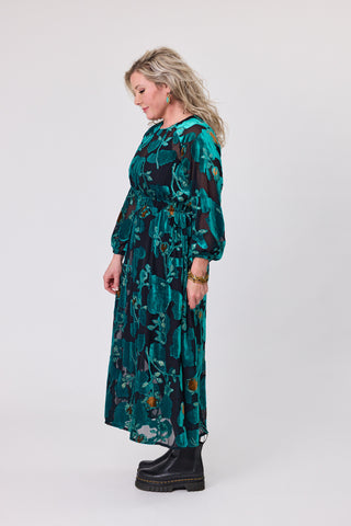 Side view of a woman wearing a teal orchid velvet dress with floral prints and a flowing skirt.