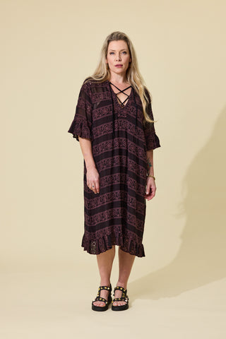 A woman modeling a dark espresso embroidery lace-up dress with ruffled sleeves and hem, paired with black platform sandals, against a solid beige studio background.