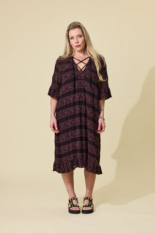 A woman modeling a dark espresso embroidery lace-up dress with ruffled sleeves and hem, paired with black platform sandals, against a solid beige studio background.