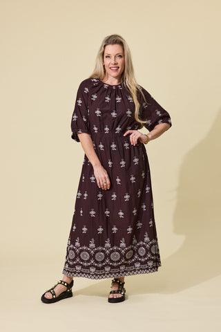 A woman modeling a dark brown bohemian-style maxi dress with white floral and geometric patterns, standing against a solid beige studio background.