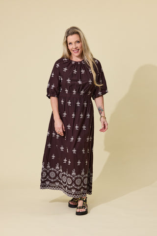A woman modeling a dark brown midi dress with a white floral and geometric pattern, standing against a solid beige studio background.