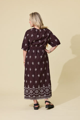A woman modeling a dark brown midi dress with a white floral block print and a circular embroidered border at the hem, shown from the back against a solid beige background.