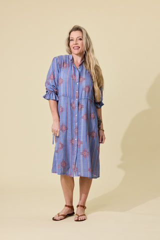 A woman modeling a blue embroidered patterned button-up shirt dress with ruffled sleeves, standing against a solid beige studio background.