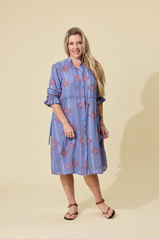 A woman modeling a blue embroidered patterned button-up shirt dress with ruffled sleeves, standing against a solid beige studio background.