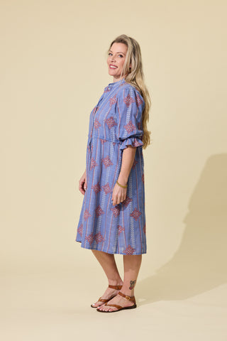 A woman modeling a blue embroidered patterned button-up shirt dress with ruffled sleeves, standing against a solid beige studio background.