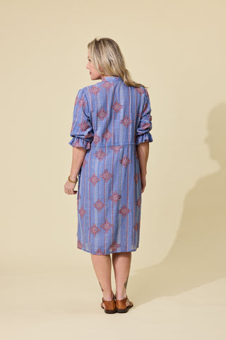 A woman modeling a blue embroidered patterned button-up shirt dress with ruffled sleeves, standing against a solid beige studio background.