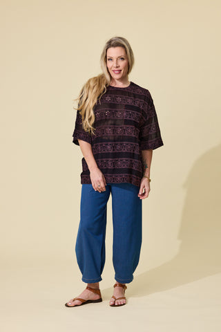 A woman modelling a espresso embroidered short-sleeve top and blue barrel jeans against a solid beige studio background.