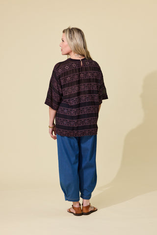 A woman modelling a espresso embroidered short-sleeve top and blue barrel jeans against a solid beige studio background.