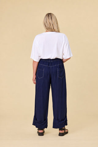 A person shown from the back wearing indigo sahara barrel-leg pants with cuffs, a white short-sleeve top, and black sandals, against a plain beige studio background.