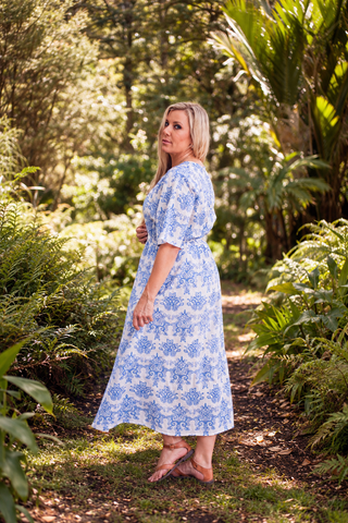 A woman wearing a blue and white embroidered Devotion midi dress stands on a sunlit garden path surrounded by lush green foliage. libertine the label