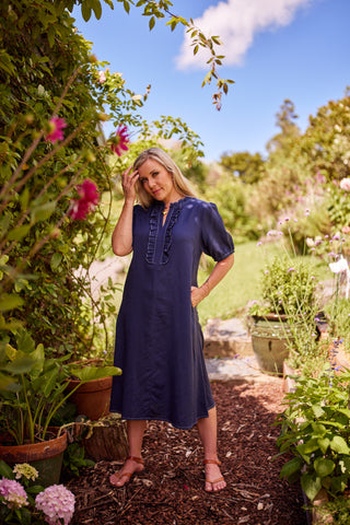 A woman wearing a indigo blue midi dress with ruffle front and white topstiching stands on a garden path surrounded by plants, flowers, and trees under a blue sky.