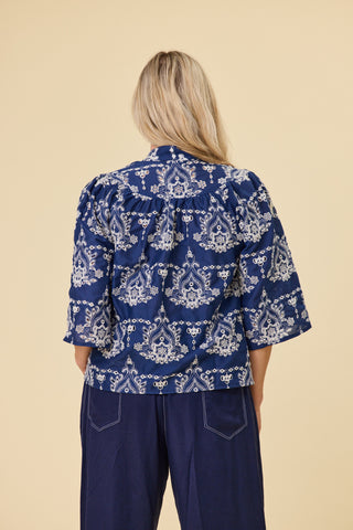 A woman modeling a indigo blue blouse with a ivory embroidery pattern, featuring a keyhole tie neckline and elbow-length sleeves, paired with indigo sahara pants against a solid beige background.