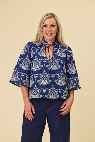 A woman modeling a indigo blue blouse with a ivory embroidery pattern, featuring a keyhole tie neckline and elbow-length sleeves, paired with indigo sahara pants against a solid beige background.