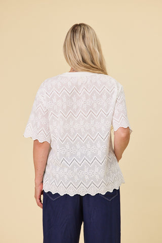A woman modeling a coconut coloured embroidery luna lace top with a scalloped hem, paired with indigo blue barrel-leg sahara pants and black sandals, against a solid beige studio background.