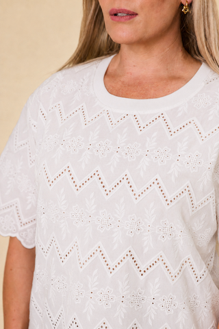 A woman modeling a coconut coloured embroidery luna lace top with a scalloped hem, paired with indigo blue barrel-leg sahara pants and black sandals, against a solid beige studio background.