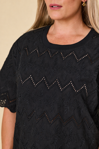 A woman modeling a black short-sleeve luna top and barrel-leg panther-print sahara pants with black sandals, standing against a solid beige studio background.