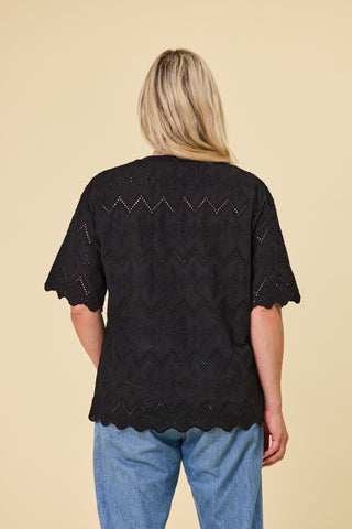 A woman viewed from the back wearing a black short-sleeve embroidery luna top with a scalloped hem and blue jeans, standing against a solid beige background.
