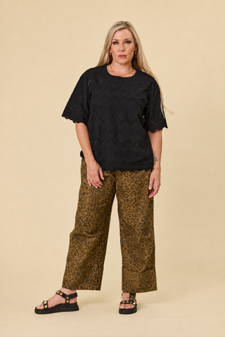 A woman modeling a black short-sleeve luna top and barrel-leg panther-print sahara pants with black sandals, standing against a solid beige studio background.