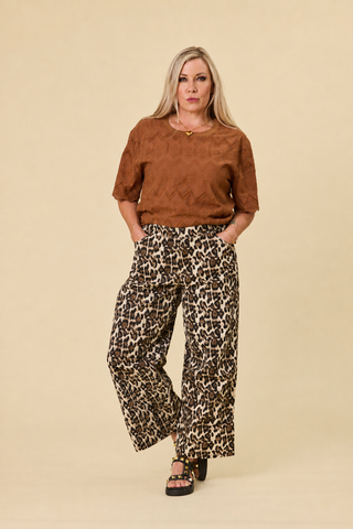 A woman modeling a hazelnut luna embroidered top and leopard print Sahara barrel jeans against a solid beige studio background.