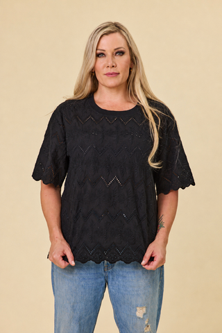 A woman modeling a black short-sleeve luna top and jeans, standing against a solid beige studio background.