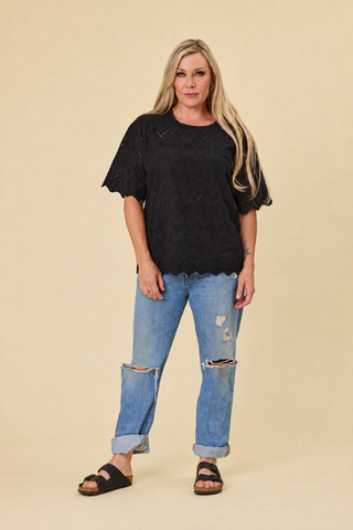 A woman modeling a black short-sleeve luna broiderie top with scalloped edges, paired with distressed blue jeans and black slide sandals, against a solid beige studio background.