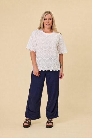 A woman modeling a coconut coloured embroidery luna lace top with a scalloped hem, paired with indigo blue barrel-leg sahara pants and black sandals, against a solid beige studio background.
