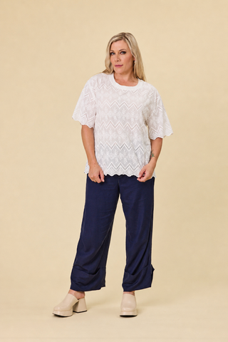 A woman modeling a coconut coloured embroidery luna lace top with a scalloped hem, paired with indigo blue barrel-leg sahara pants and black sandals, against a solid beige studio background.