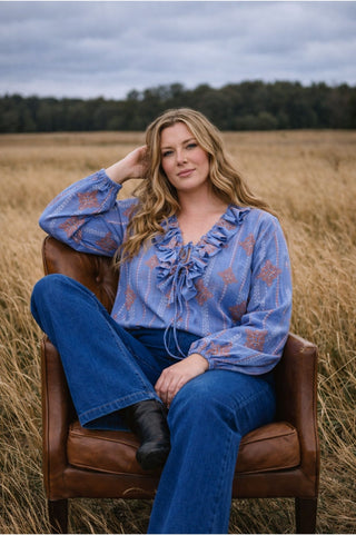 plus size Woman sitting on a brown chair in a field wearing a blue embroidered cross stitch blouse and jeans