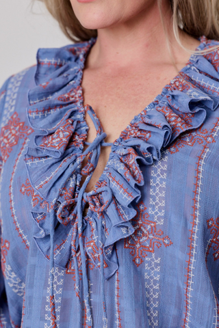 Close-up of a woman wearing a blue ruffled blouse with red and white embroidered patterns and tie details at the neckline.