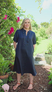 A woman wearing a indigo blue midi dress with ruffle front and white topstiching stands on a garden path surrounded by plants, flowers, and trees under a blue sky.