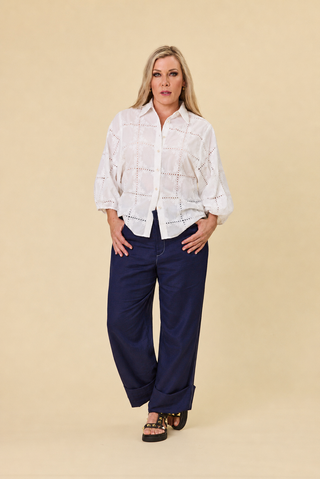 A woman modeling a white embroidered belle blouse with a keyhole neckline and three-quarter sleeves, paired with indigo sahara wide-leg barrel pants and black strappy sandals, against a solid beige studio background.