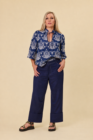 A woman modeling a blue and white embroidered devotion blouse with a keyhole neckline and three-quarter sleeves, paired with indigo sahara wide-leg barrel pants and black strappy sandals, against a solid beige studio background.