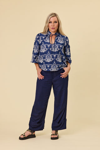 A woman modeling a indigo blue blouse with a ivory embroidery pattern, featuring a keyhole tie neckline and elbow-length sleeves, paired with indigo sahara pants against a solid beige background.