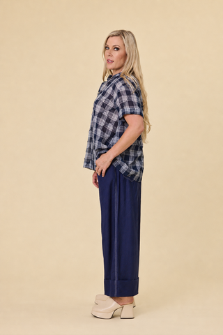 A woman modeling a blue and white plaid florence blouse , paired with indigo sahara wide-leg barrel pants, against a solid beige studio background.